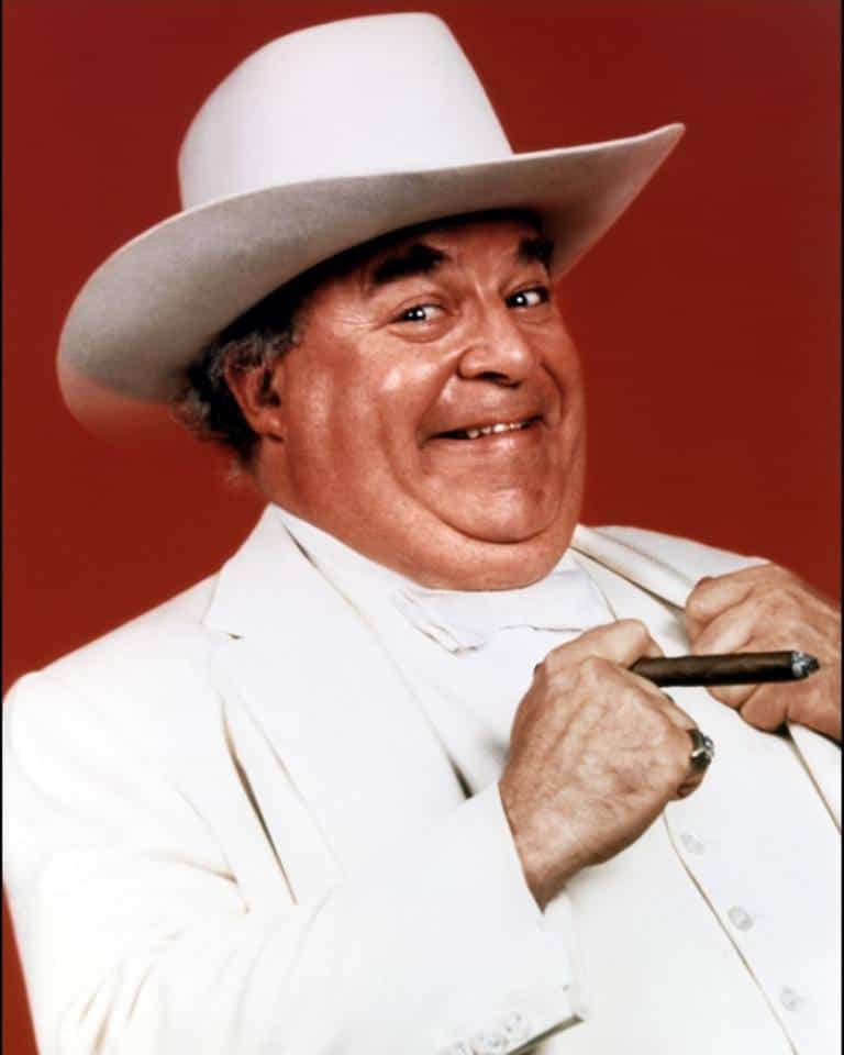 Boss hog from the Dukes of Hazzard in his white suit and hat holding a cigar with a terrible smile on his face.