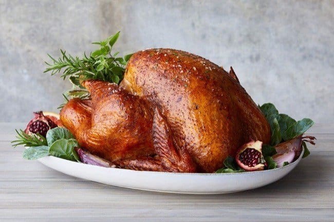 a roasted turkey surrounded by herbs and pomegranate chunks sits on a platter with a professional photo backdrop behind.