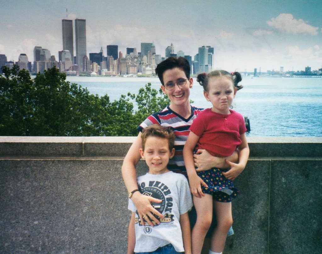 Remembering when.... 9/11 A woman in a red, white, and blue shirt stands on Liberty Island with her 5 yo son and holds her 2yo daughter. The pre-9/11 twin towers are visible across the water.