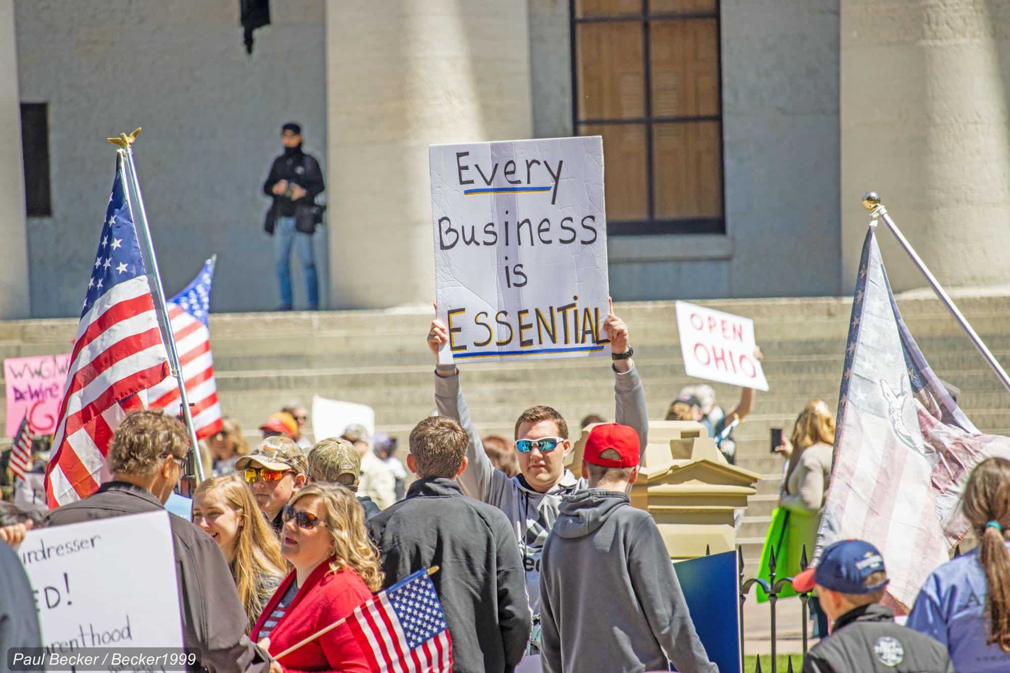 A man stands below the steps of a building with a protest sign saying 'every business is essential'.