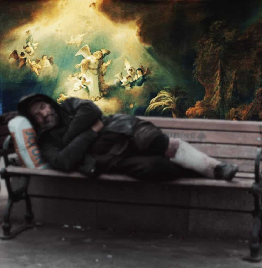 Image shows a man sleeping in the rough on an outdoor bench with a vibrant angelic scene behind him.