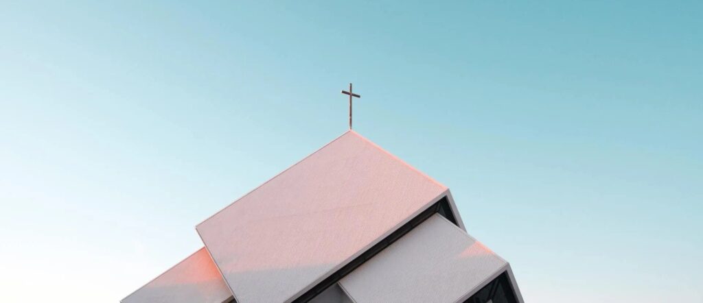 The top of a steep two level church roof is shown against a blue sky and with a cross on it's peak.