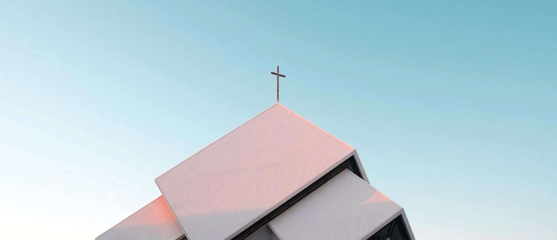 The top of a steep two level church roof is shown against a blue sky and with a cross on it's peak.
