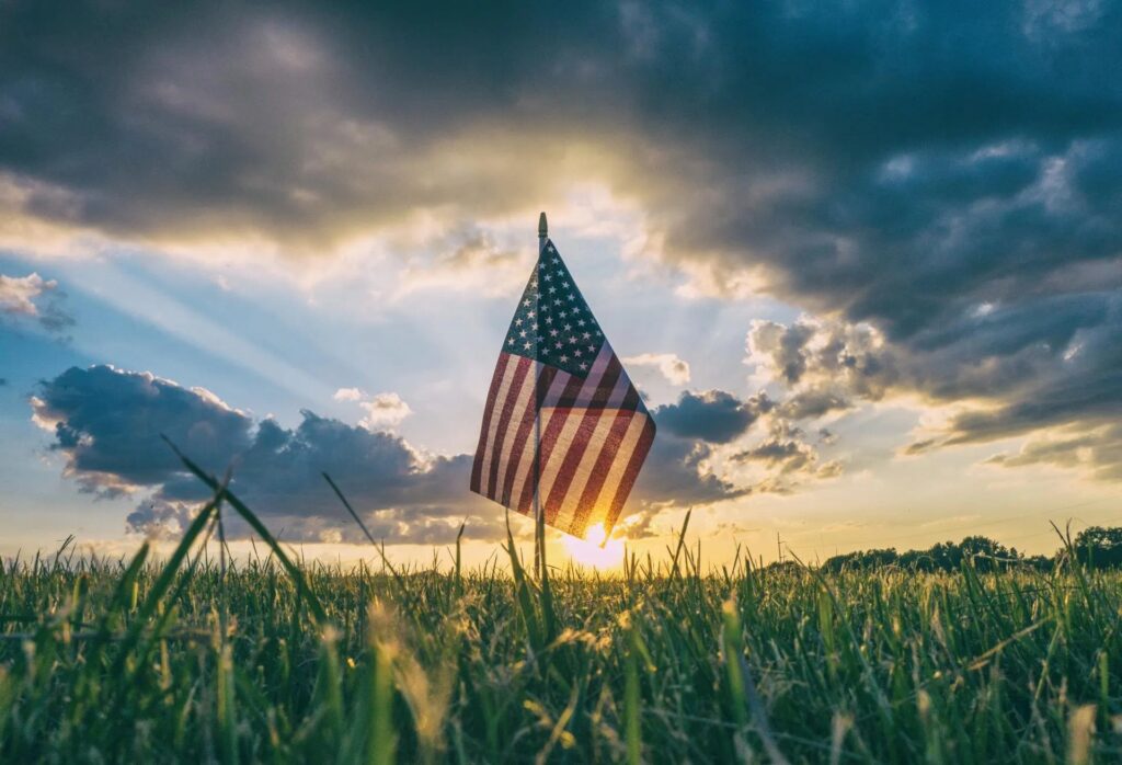 An American flag in a field with the setting sun and clouds in the background.