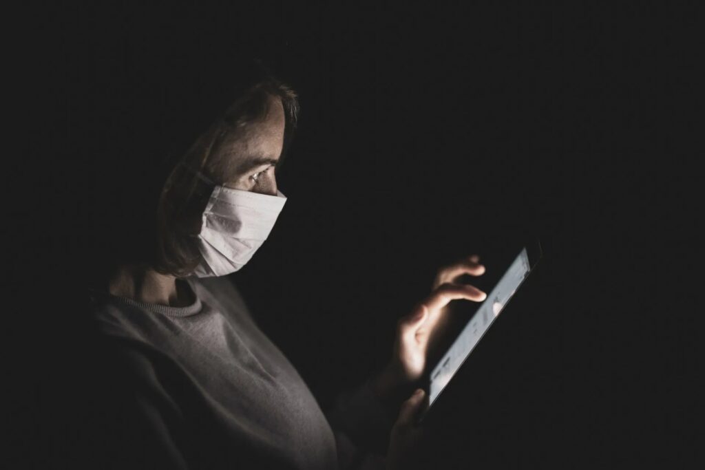 A masked person sits in a very dark room swiping at a phone.