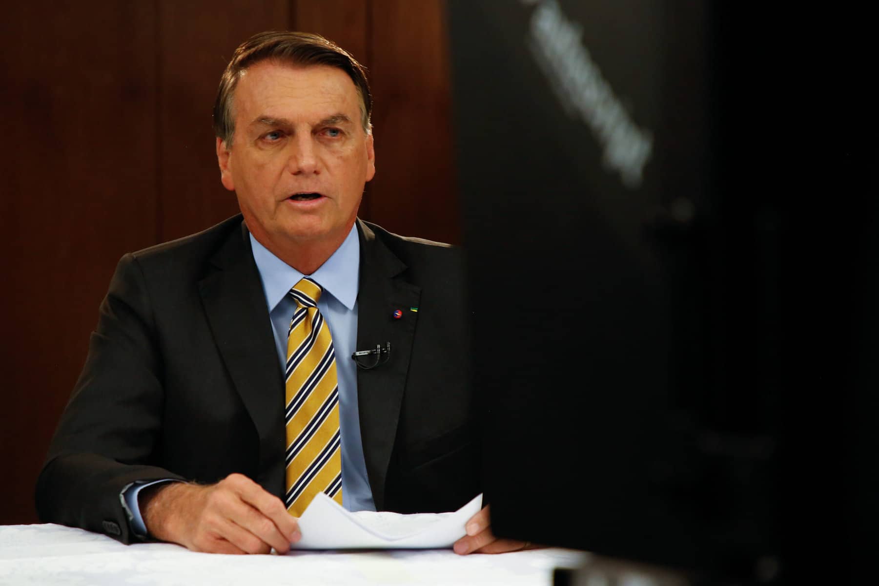 A man in a suit and tie and holding a piece of paper sits at a newscaster's desk in front of a camera.