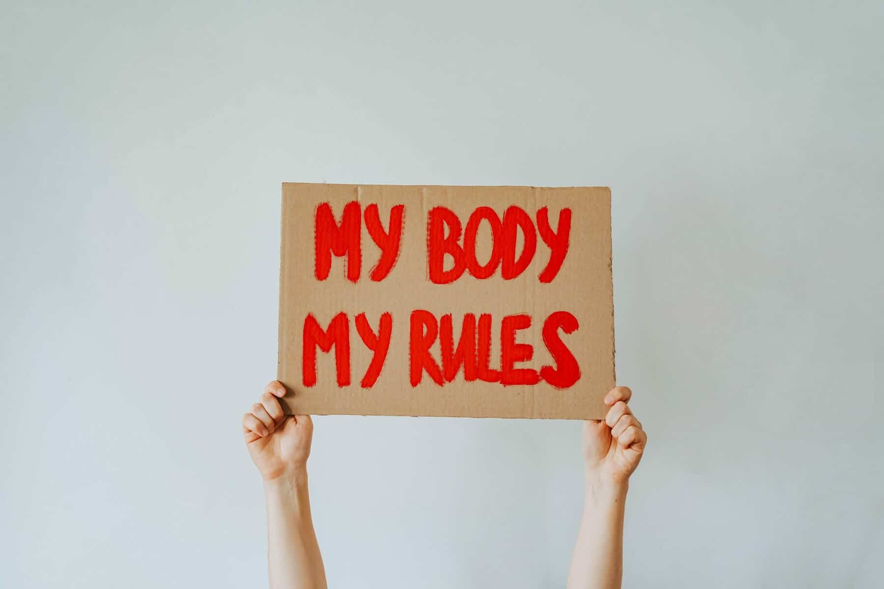 A woman's hands hold up a cardboard sign with "My body my rules" painted on it in red.