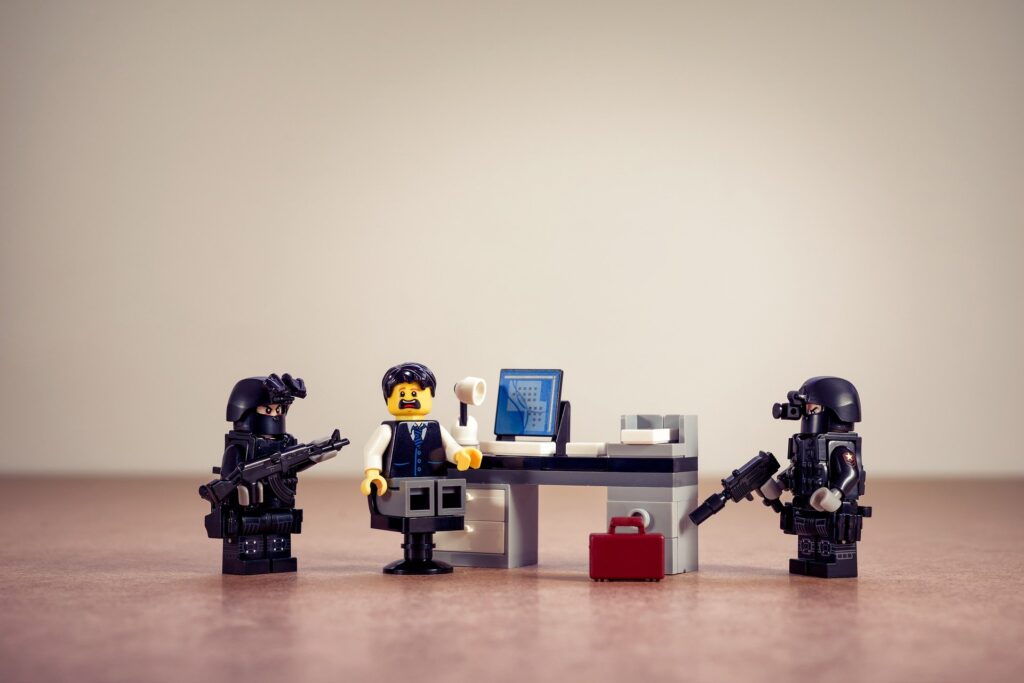 Pro-Life Christian’s home raided by FBI, guns drawn A Lego minifig sits in a chair at a desk with a surprised look. To each side is a minifig dressed in police-type gear with long guns.