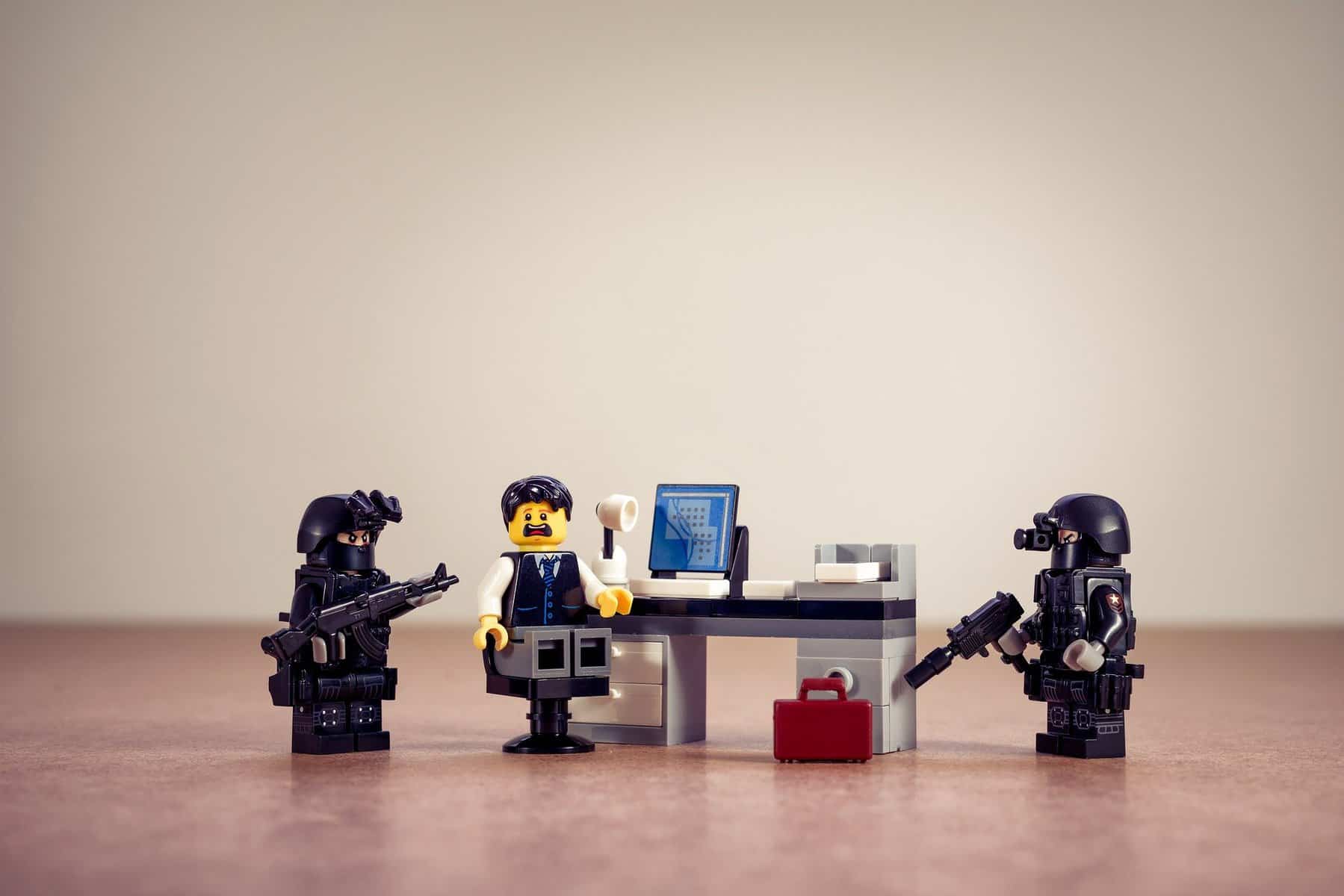A Lego minifig sits in a chair at a desk with a surprised look. To each side is a minifig dressed in police-type gear with long guns.