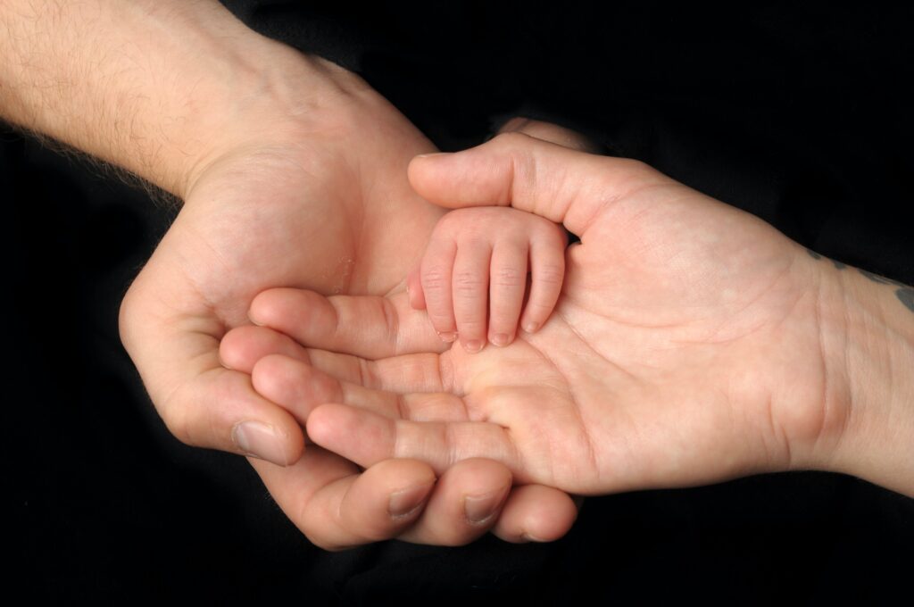 Beloved Child of God Two Caucasian adult hands, one in the other, with a baby's hand in them.