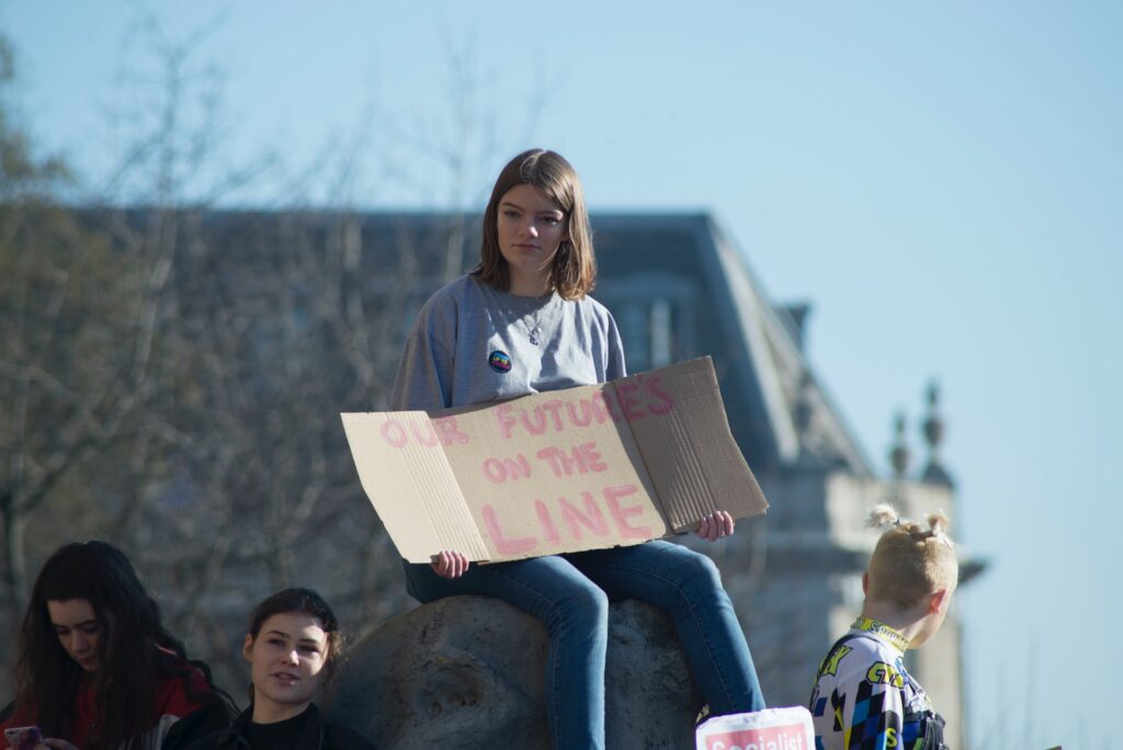 A teen girl sits on a boulder holding a cardboard sign that says in red "our future's on the line".