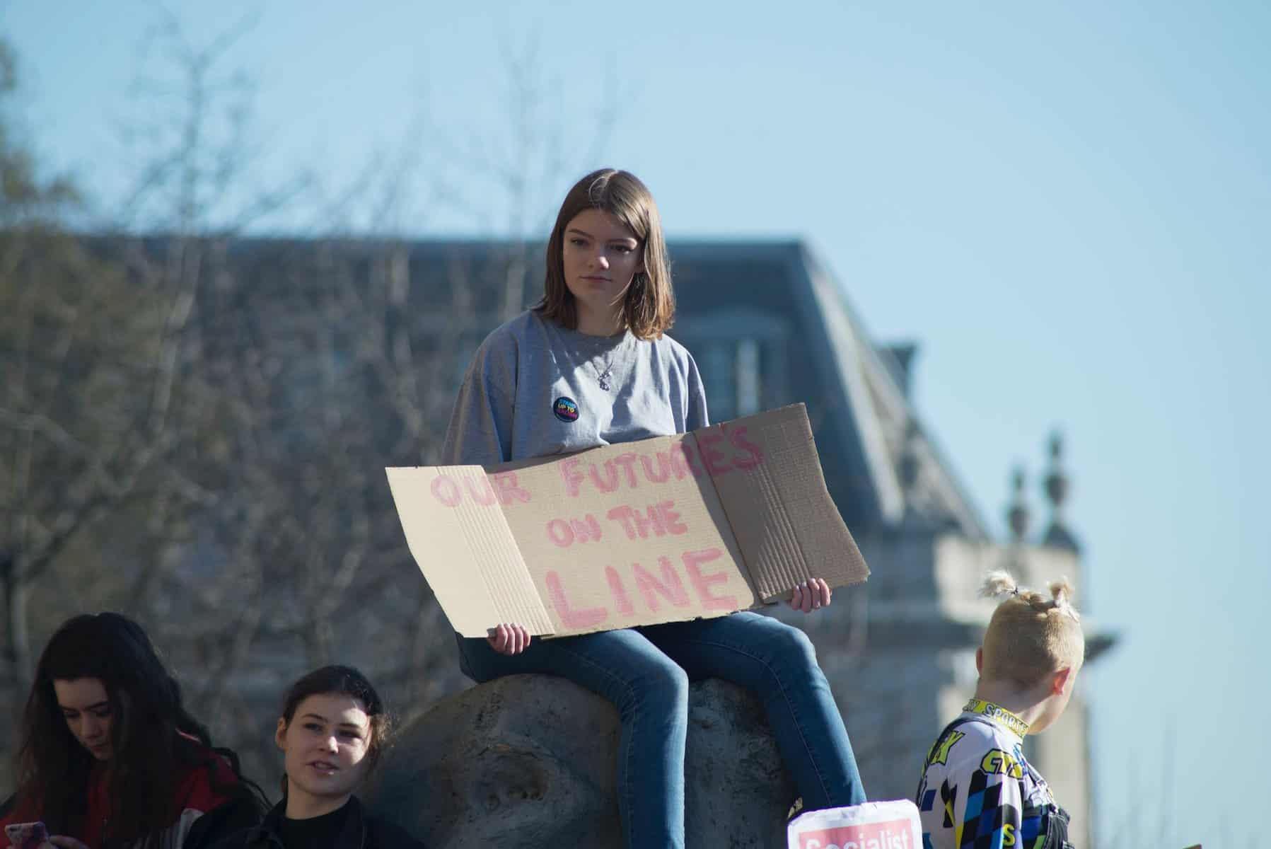 A teen girl sits on a boulder holding a cardboard sign that says in red "our future's on the line".