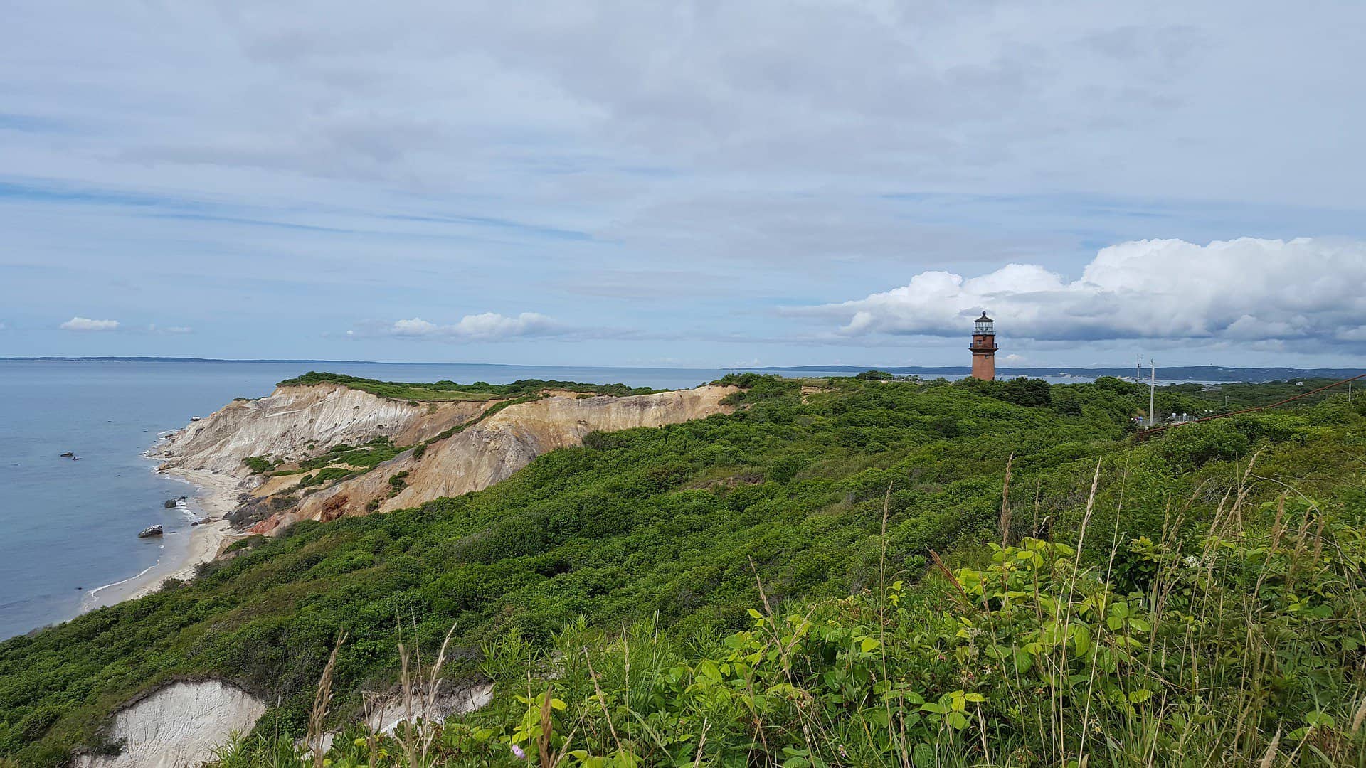 A lighthouse sits in the distance on a sandy-looking cliff surrounded by vegetation and the ocean.