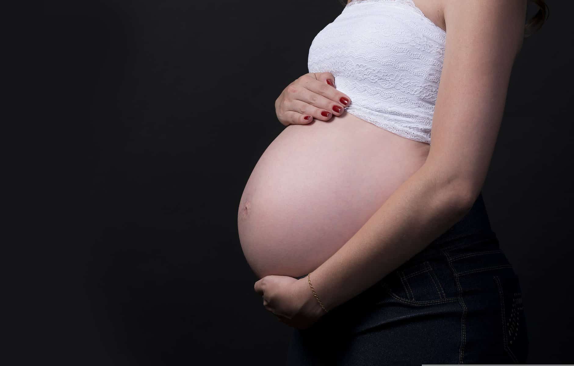 A heavily pregnant woman in jeans and a white tube top is only seen from the hips to the neck holds her bare belly in a studio photo session