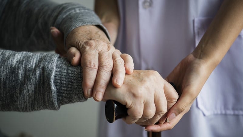 a woman medical professional is seen at torso level helping an elderly person who's hands are on a cane
