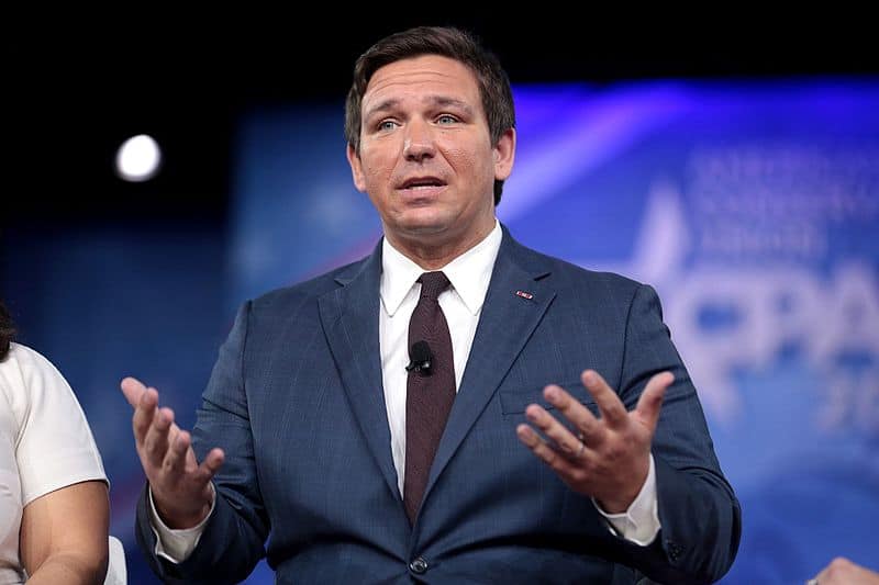 Florida Governor, Ron DeSantis, stands in front of a projected image with his hands up in the air, palms up while speaking