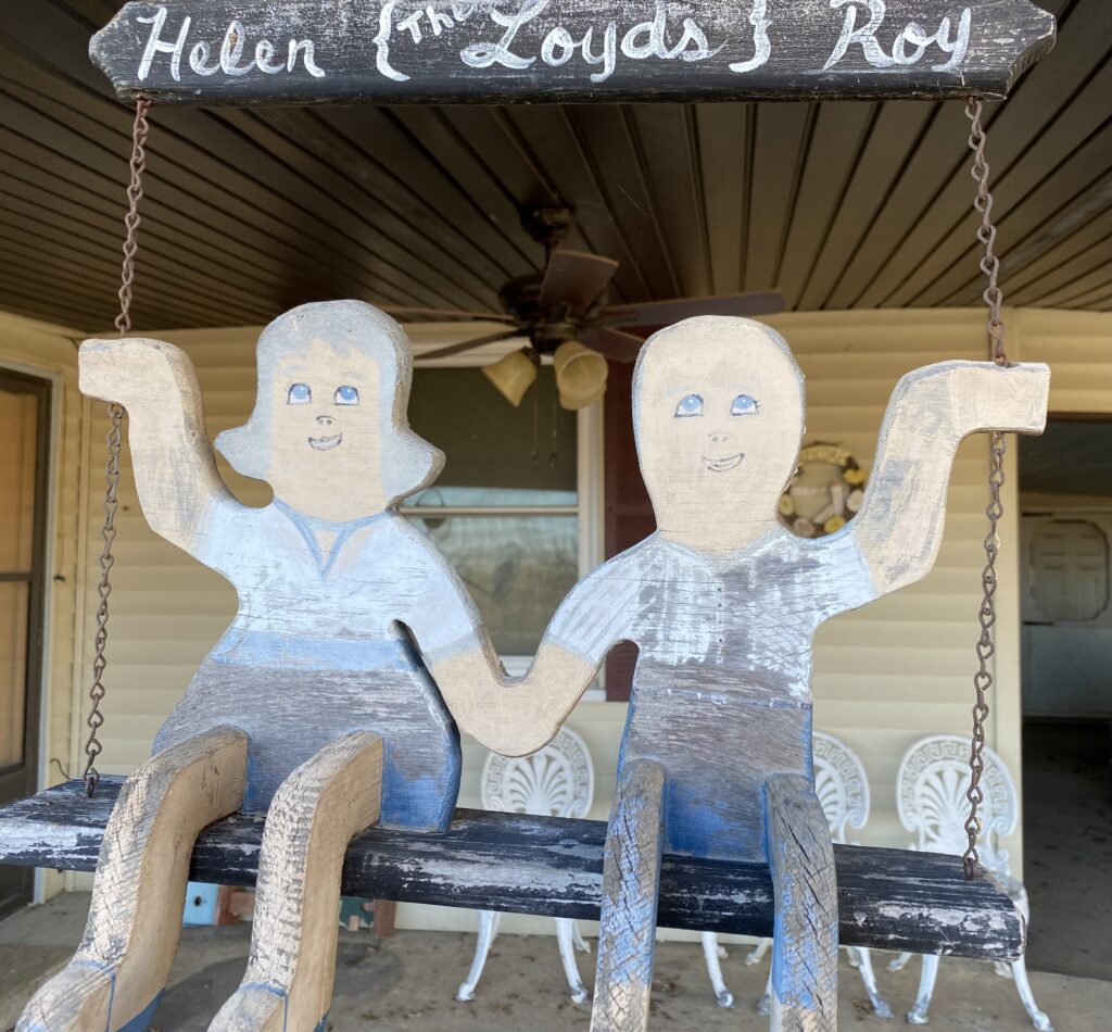 The Perspective That Informs My Gratitude male and female wood cut outs sit on a wooden 'bench' holding hands with a sign above saying Helen and Roy, the Loyd's