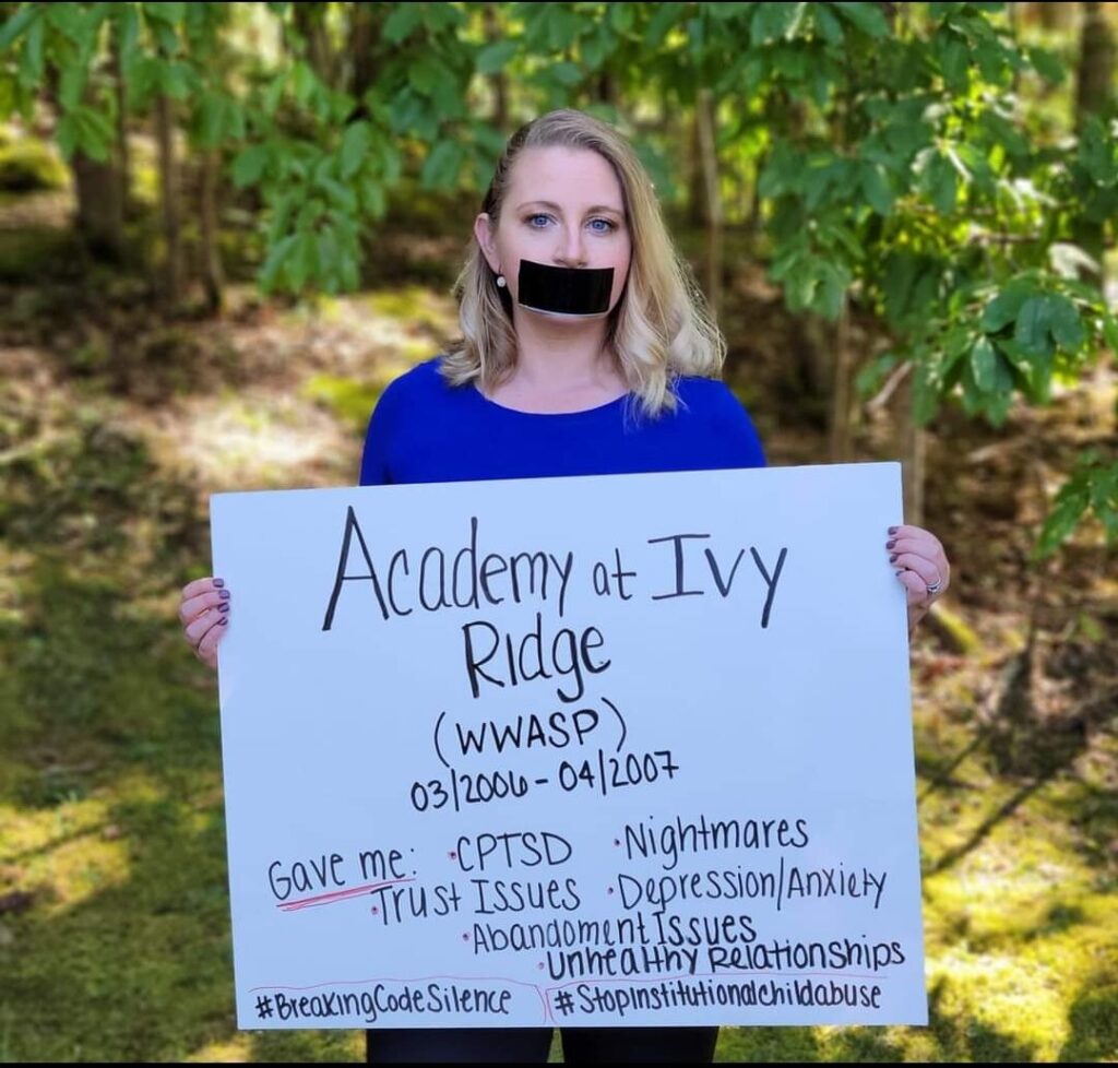 a thin young woman with blond hair holds a sign in front of her with problems she's faced due to attendance at Academy of Ivy Ridge. Her mouth is covered with black tape.