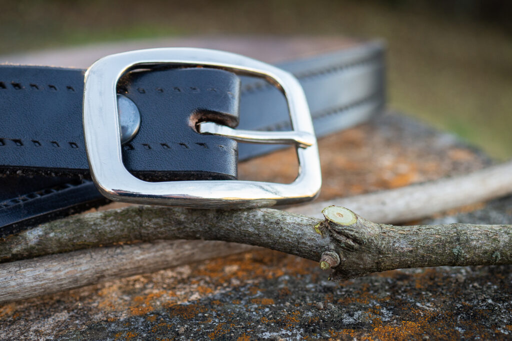 Is Spanking Christlike? In a close up, the silver buckle of a black leather belt sits on a couple twigs, one with a pruning cut. All items sit on old weathered concrete.