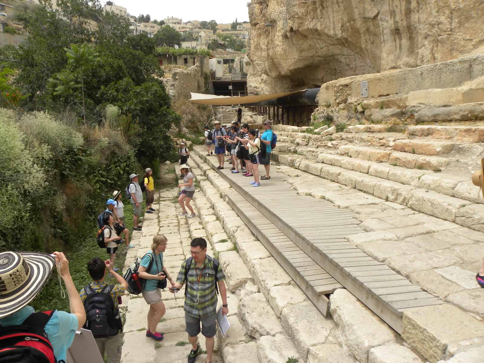 Tourists with hats and backpacks walk along the steps of the Pool of Siloam and take photos.