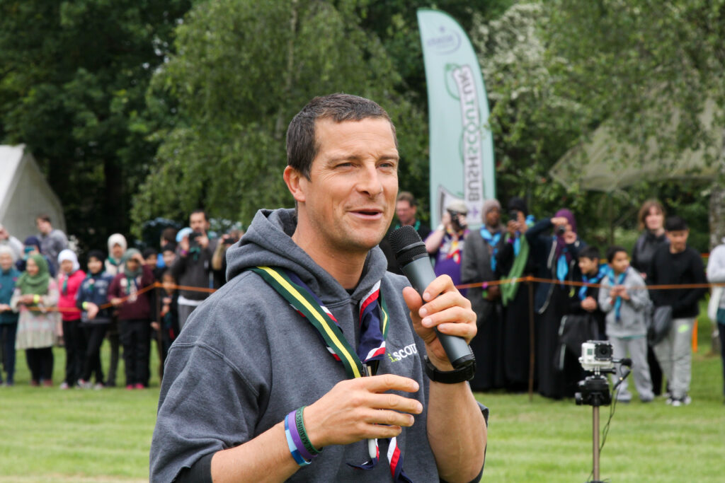 Bear Grylls says modern Western church doesn’t represent what Jesus would want Bear Grylls is shown in casual clothes holding a microphone and speaking at an outdoor event.