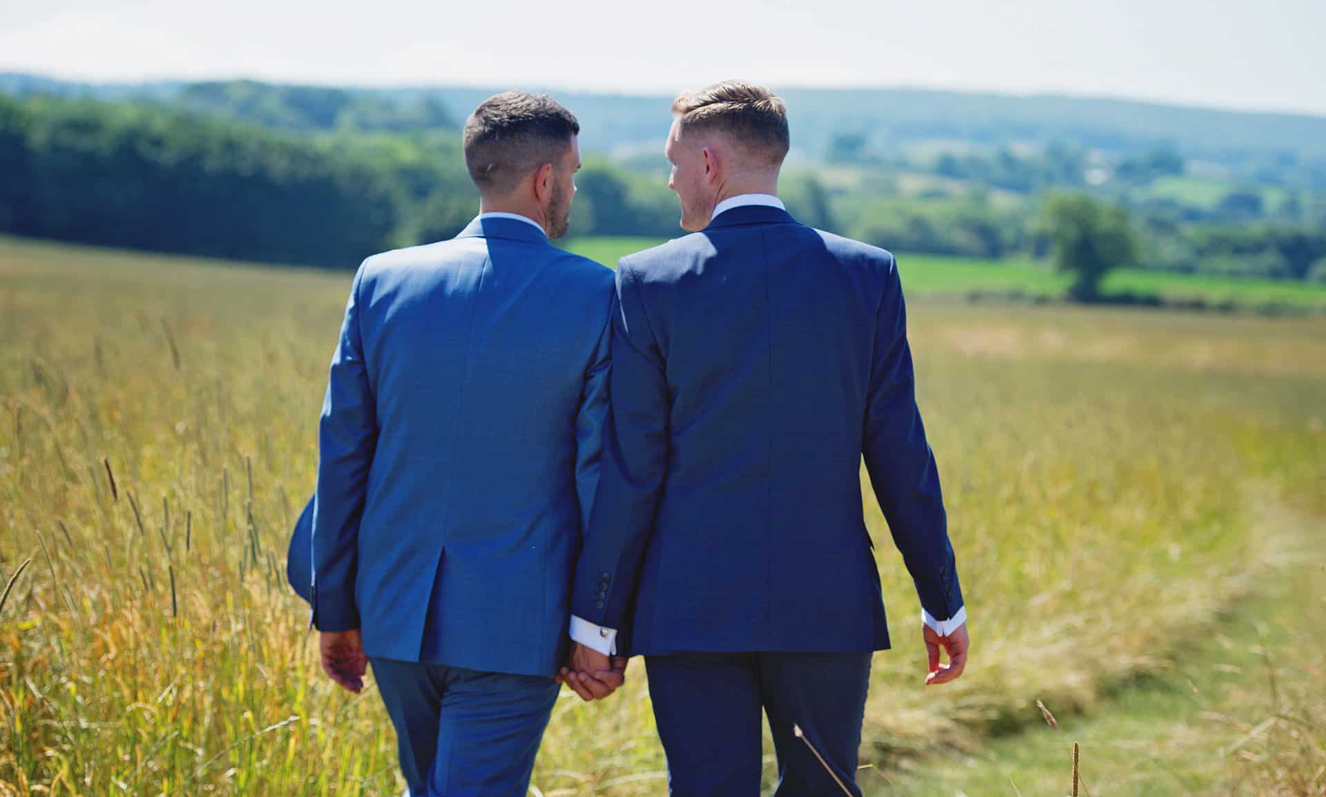 Two men dressed in blue suits hold hands and walk away from camera in a pastoral setting.