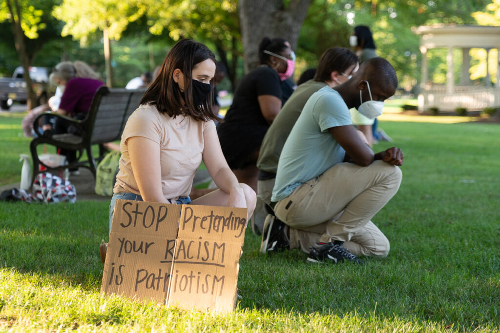 A Call for Christians: Challenge Hell's Native Tongue by Telling the Truth About American History A young woman kneels at a protest behind a cardboard sign on which is written "Stop pretending that your racism is patriotism."