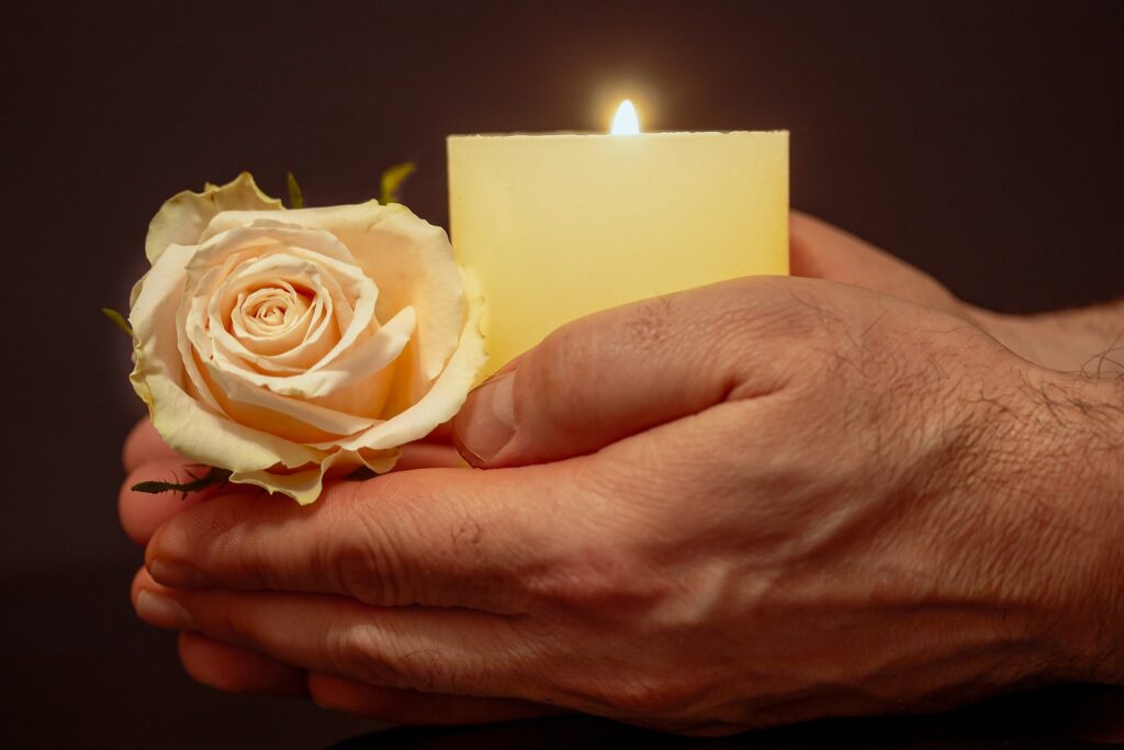 Church of England seeks new home for remains of Anglo-Saxon saint A Caucasian man's hands are shown holding a lit candle in a votive and and white rose.