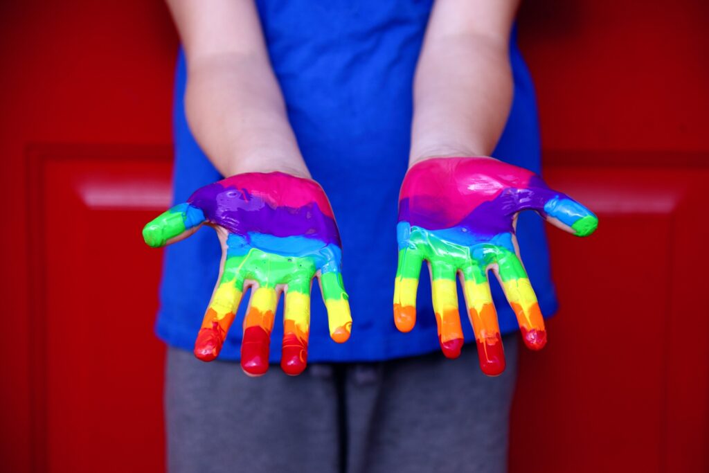Megachurch pastor to host conference for LGBTQ children’s parents A child stands in front of a red garage door in a blue t-shirt with palms up and painted in rainbow colors.