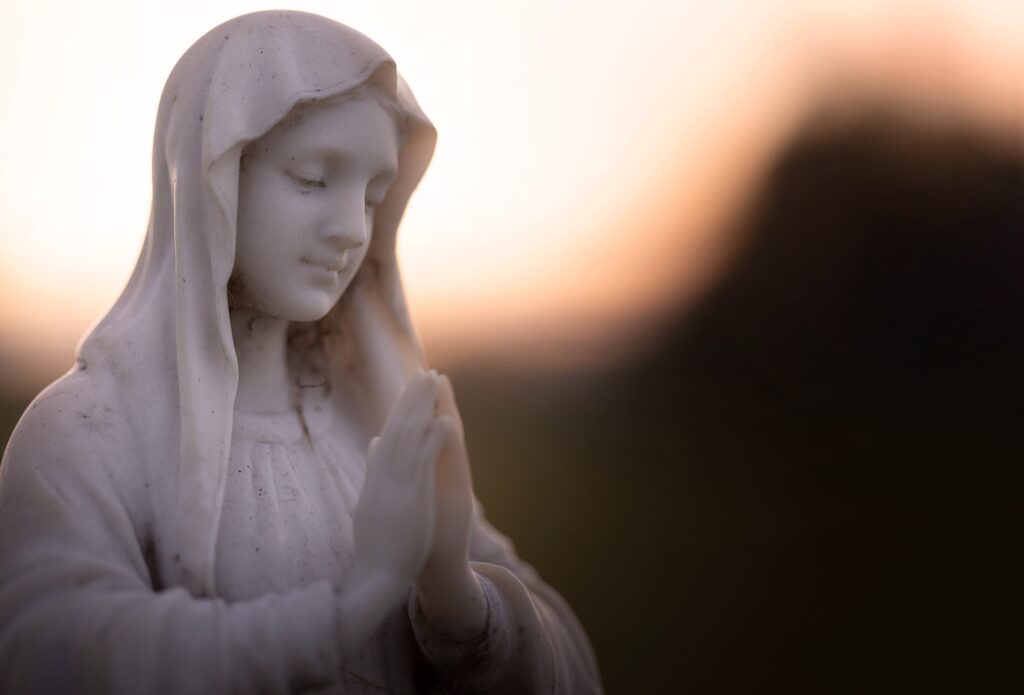 A statue of a woman with prayerful hands is shown in soft lighting.