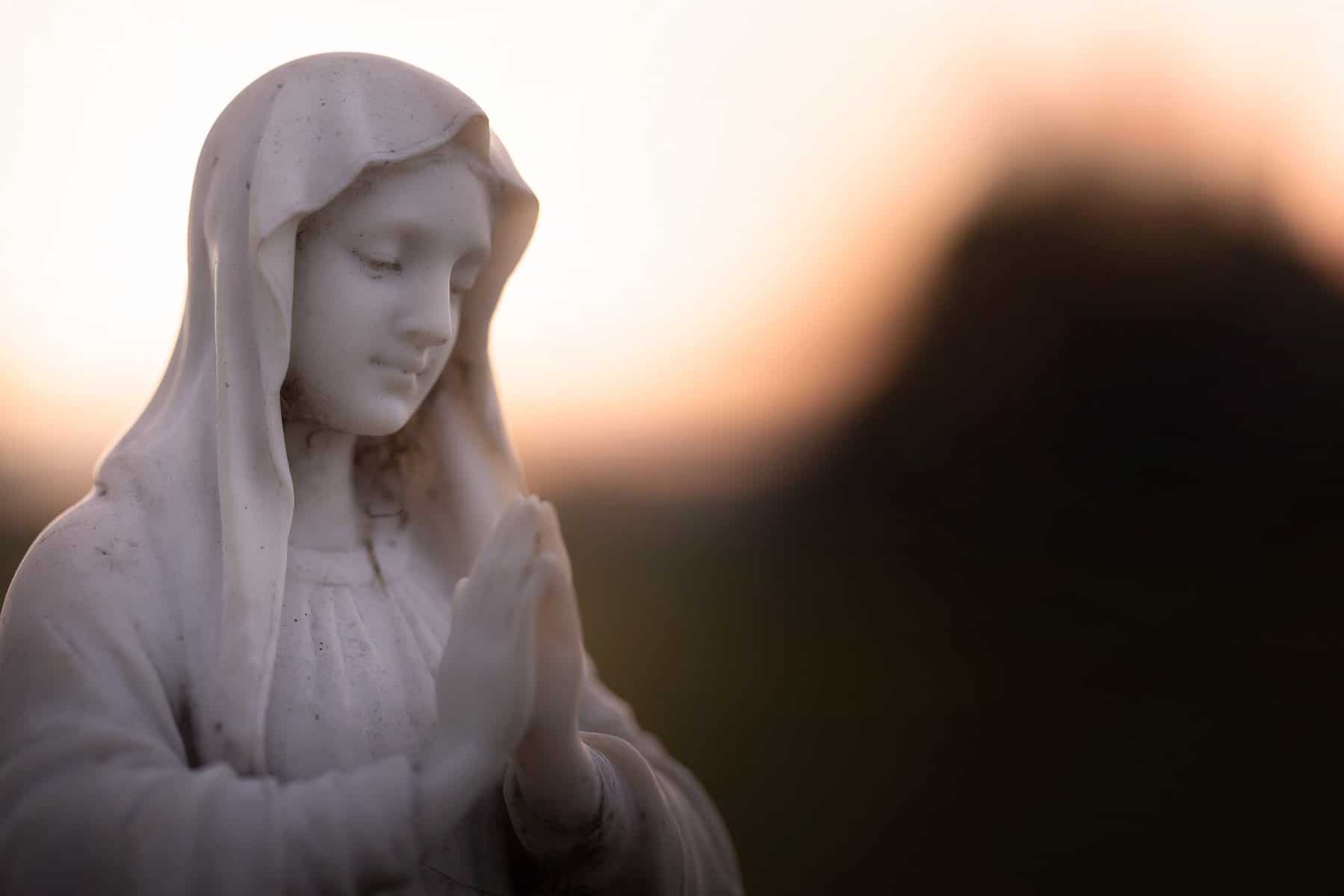 A statue of a woman with prayerful hands is shown in soft lighting.