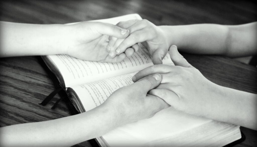 Baylor University holds 72-hour prayer event Black and white image shows two people's hands a forearms. The people are holding hands resting on a bible that is open on a table top.