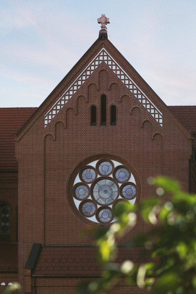 A portion of the front of a dark red brick church is shown with a stained glass rose window on the face and cross on top of the roof peak.