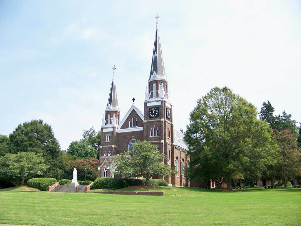 Belmont_Abbey_Historic_District A red stone church with white stone accents and steep, pointed spires with green grass shown in the foreground.