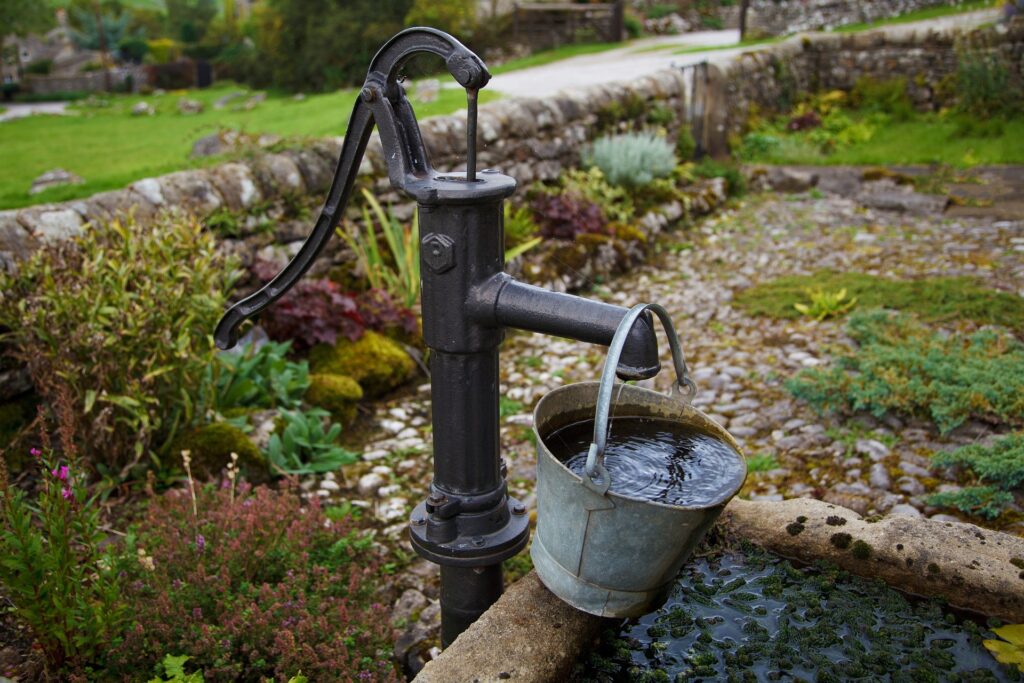 A metal bucket with water in it hangs from an antique hand water pump.