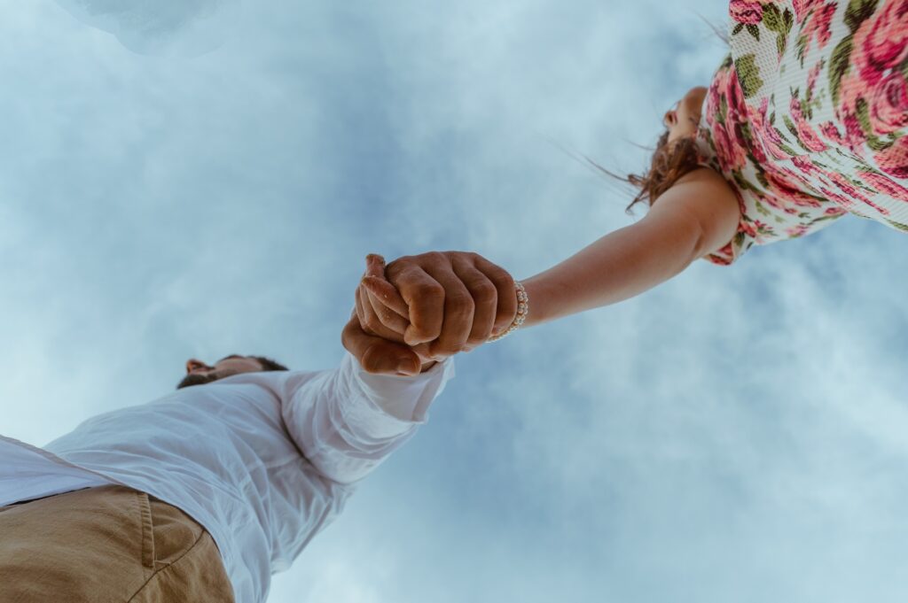 A man and woman's clasp hands are shown from directly underneath with a slightly cloudy sky above and functioning as the background.