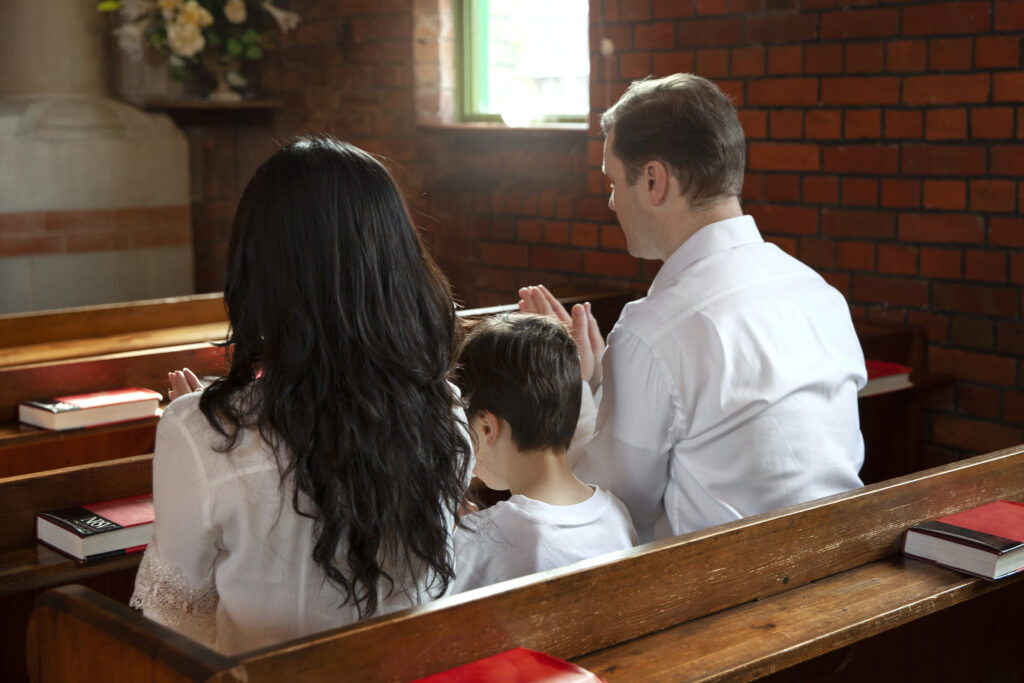 A mother, child, and father are seen in a pew from the back and in that order from a church aisle.