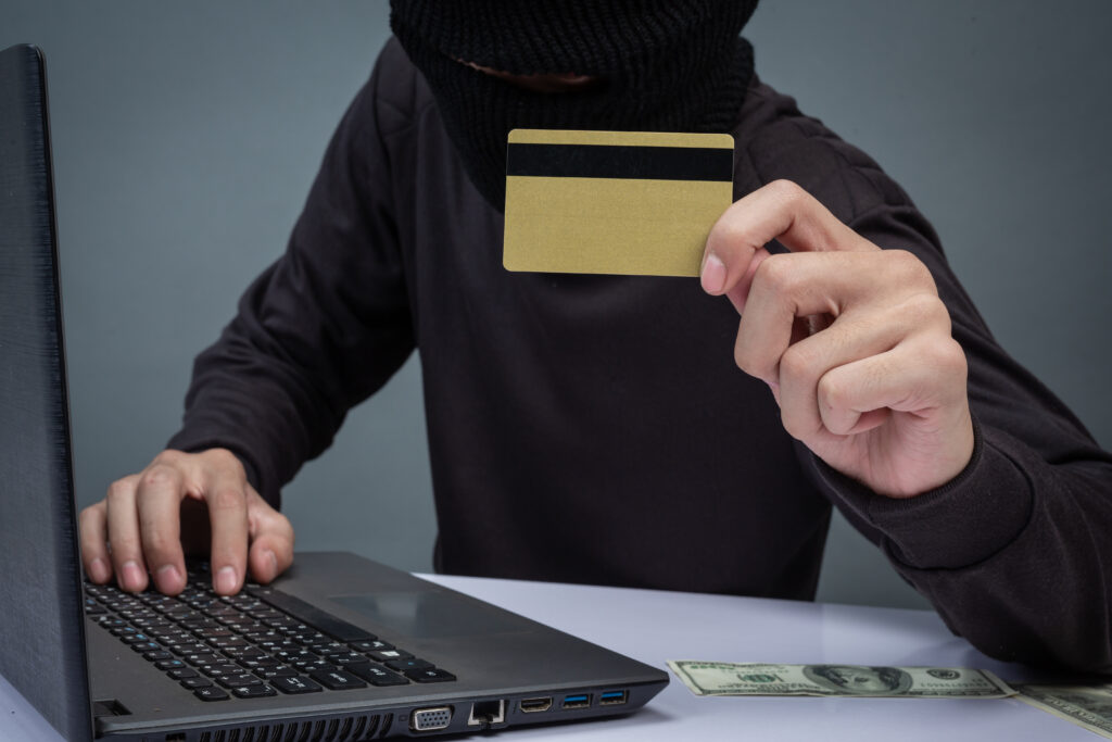 Virtual scams reach the Diocese of York A person disguised in black clothing and a mask is seen as a computer with paper money at their elbow and holding a credit card.