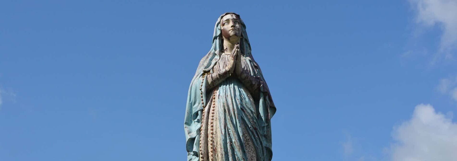A bronze statue of the virgin Mary is seen from below and against a blue sky.