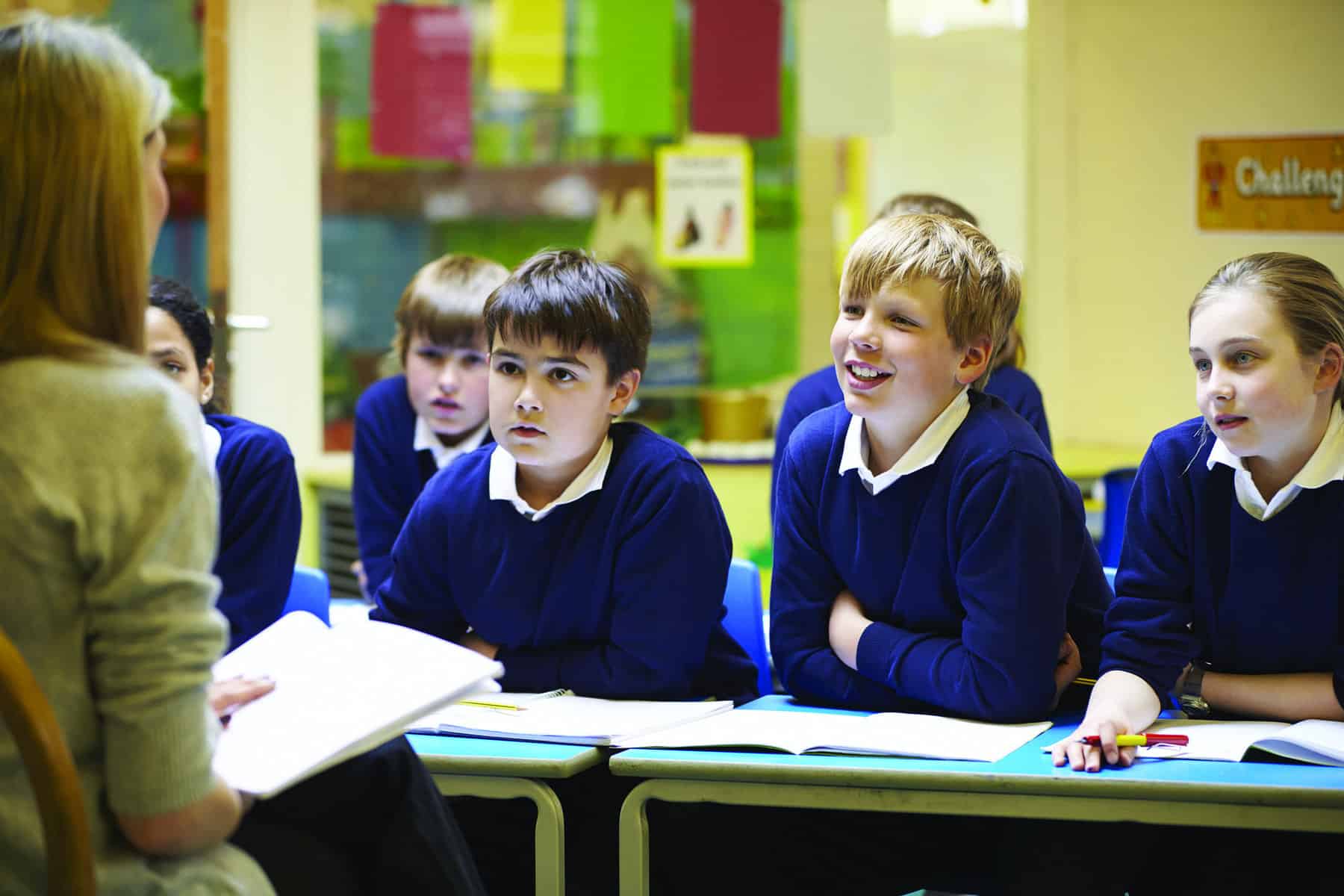 Three children are clearly seen facing the camera wearing white shirts and blue sweaters and leaning on a school table. The teacher is mostly out of frame.