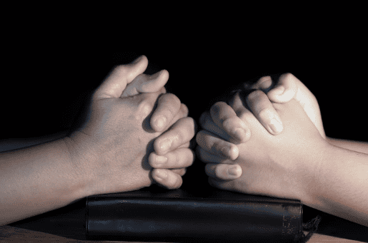 Two pairs of hands are clasped in prayer on top of a bible in this black and white image.
