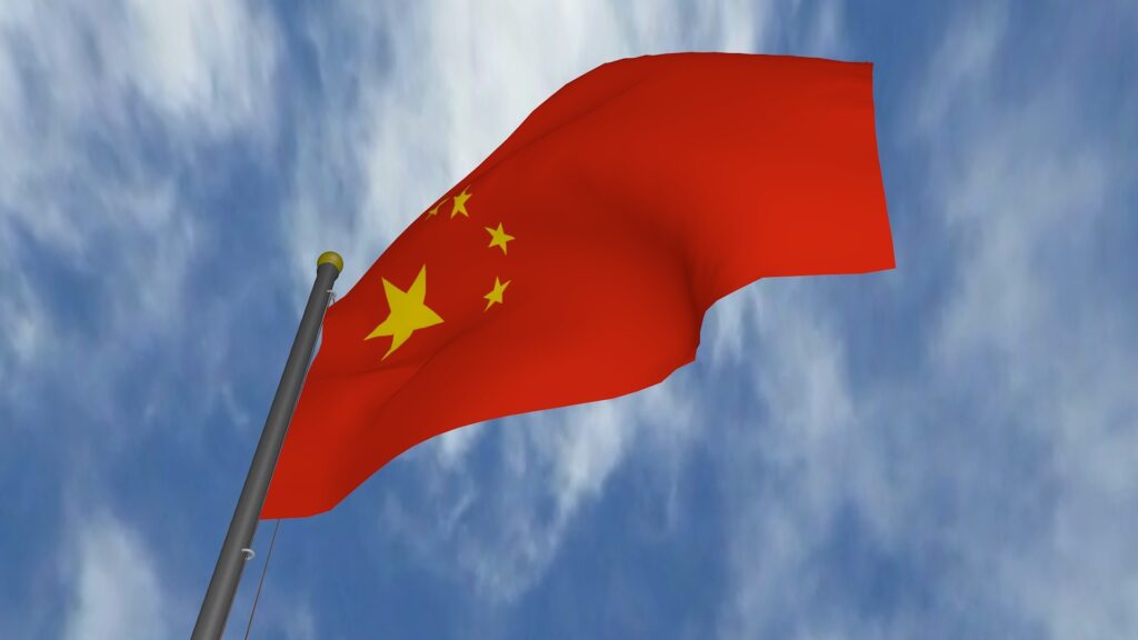A red Chinese flag on a pole is seen from below with a blue sky and thin white clouds above.