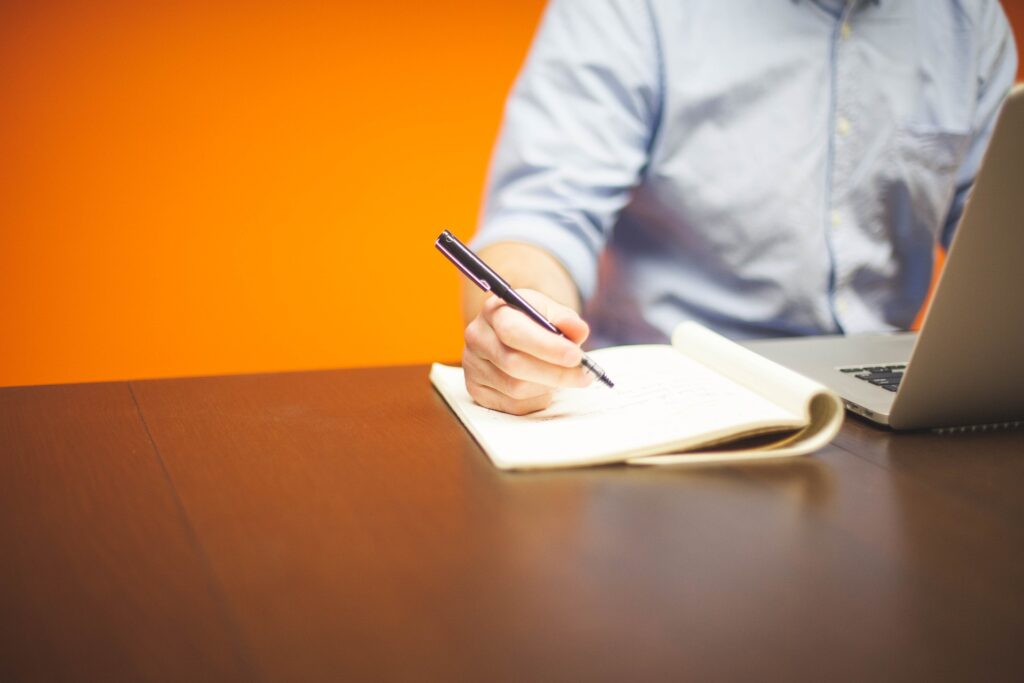 UK government should enhance its comprehension of faith: Bloom report The torso of a man is shown sitting at a computer and writing on a pad of paper against an orange background.