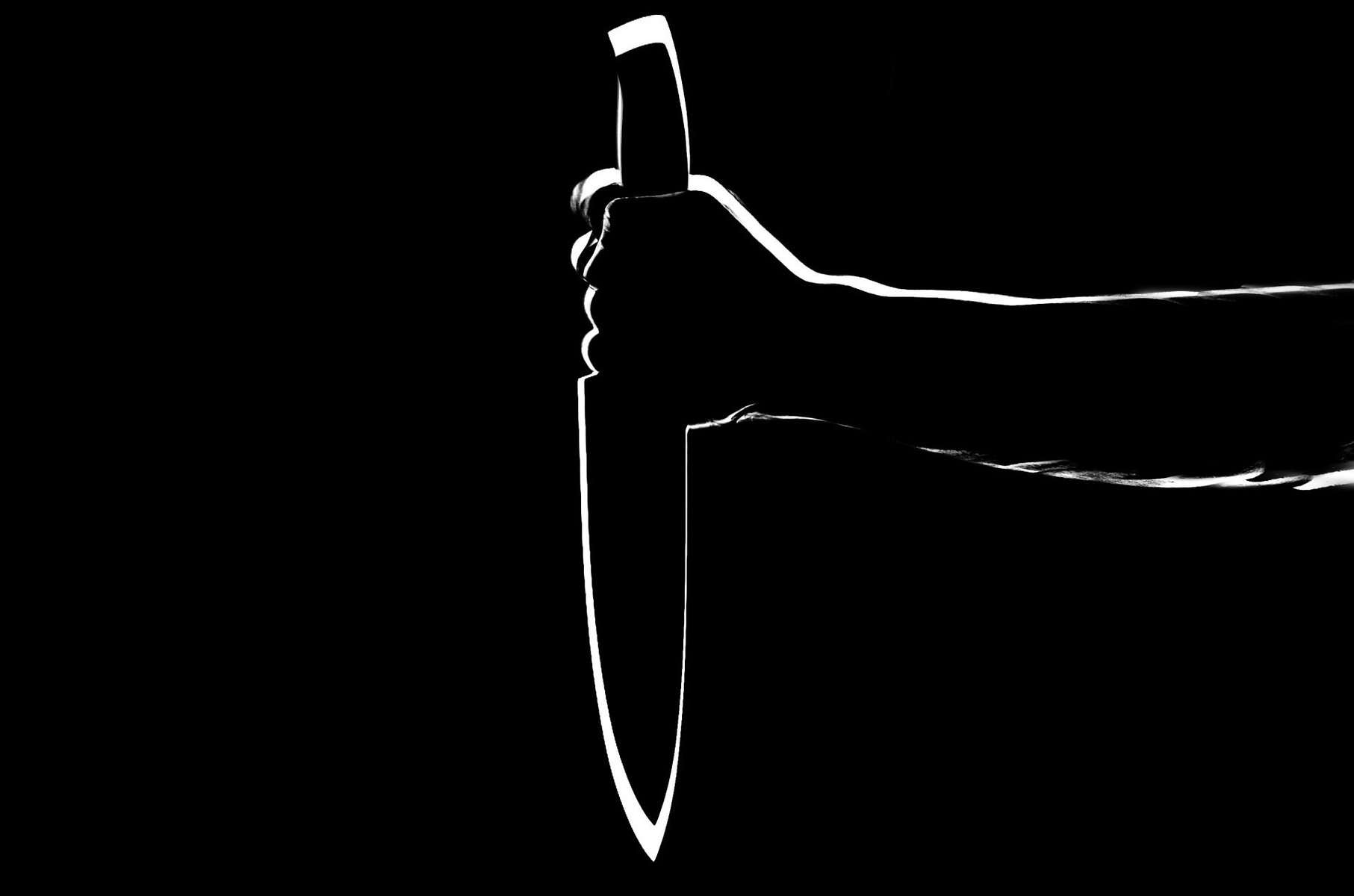 In this dark black and white image, an arm is extended holdonga knife ponted downward with blade facing away from the holder. The image has a strong rim light.