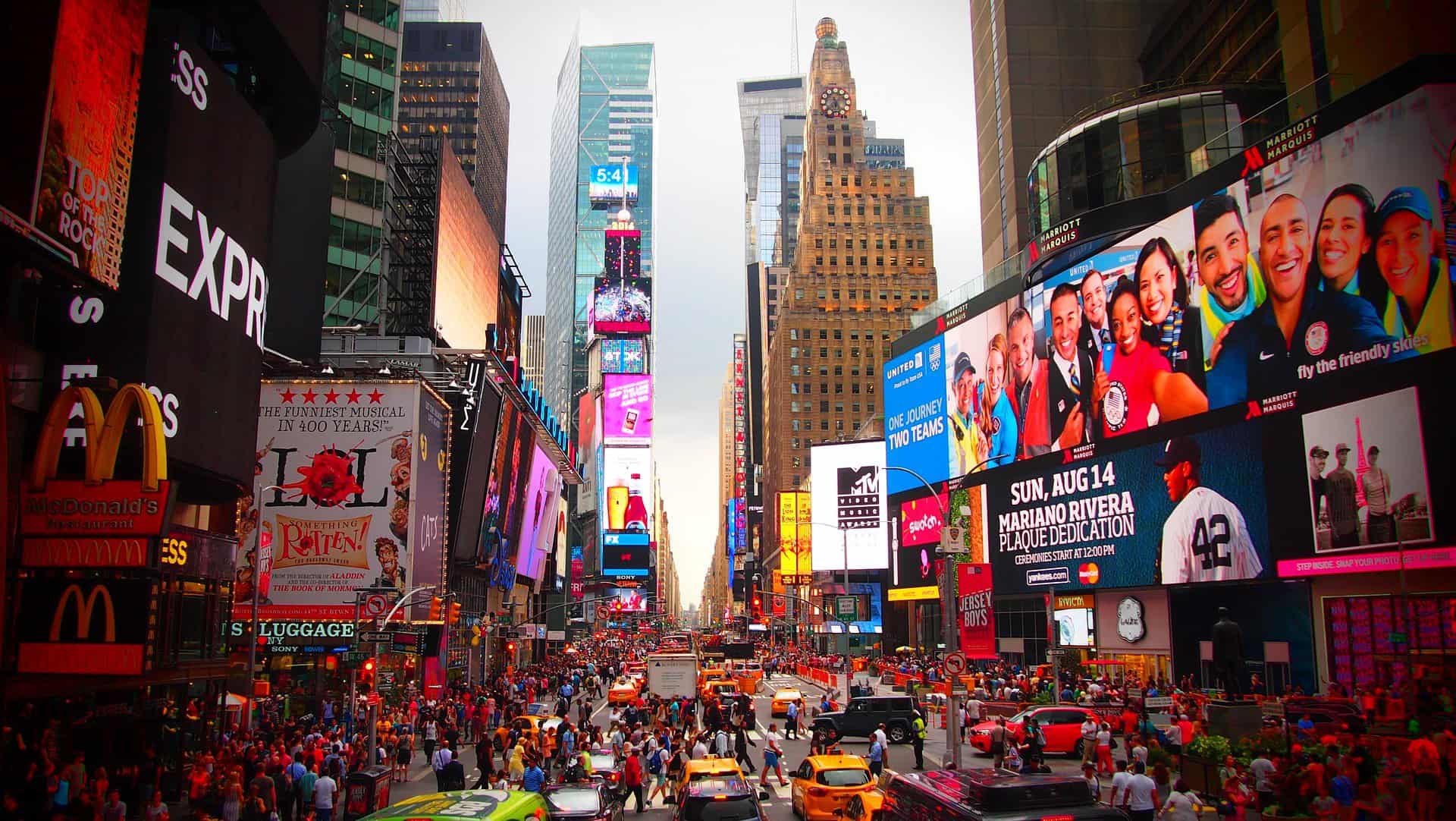Times Square is seen from a couple stories high filled with people and jumbotrons.