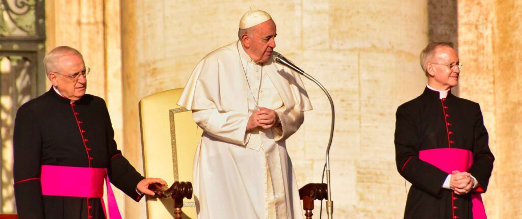 Pope Francis stands by his chair flanked by two attendants.