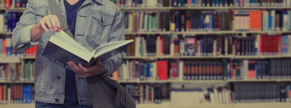 A man is seen from the shoulders to thighs leafing through a book with library shelves full of books behind him.