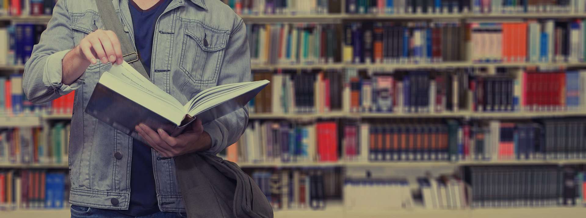 A man is seen from the shoulders to thighs leafing through a book with library shelves full of books behind him.