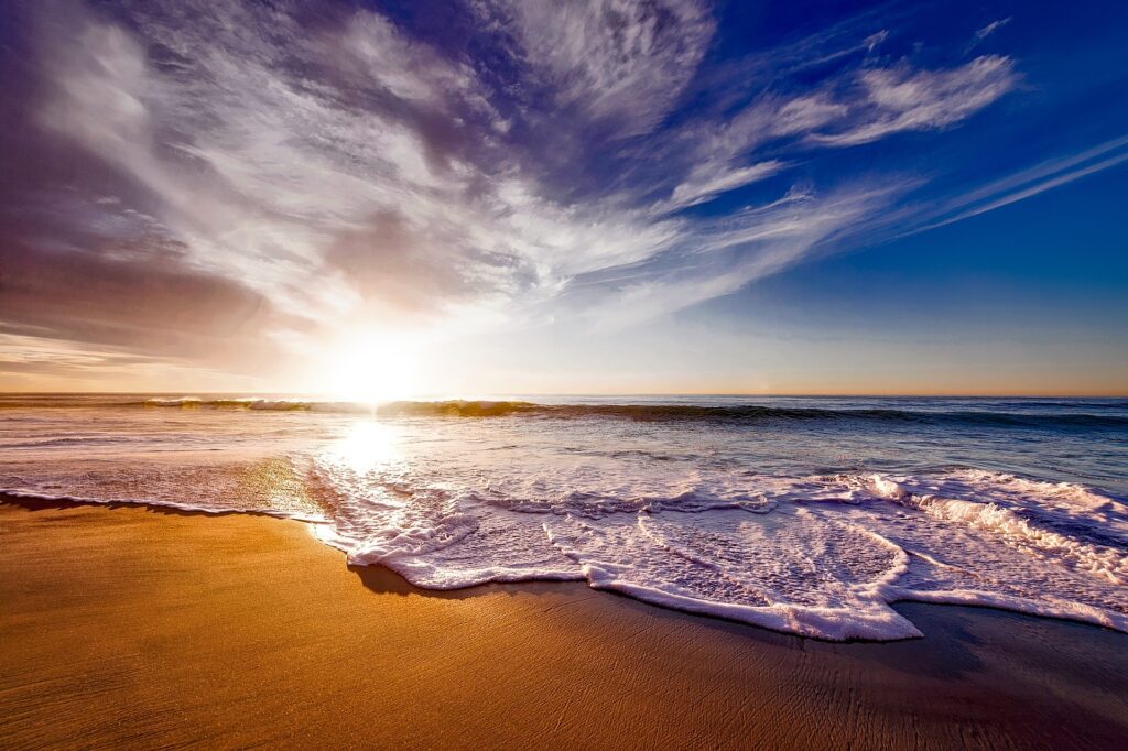 The sun is shown setting over the ocean by a camera held very low and waves are rolling over a sandy beach.