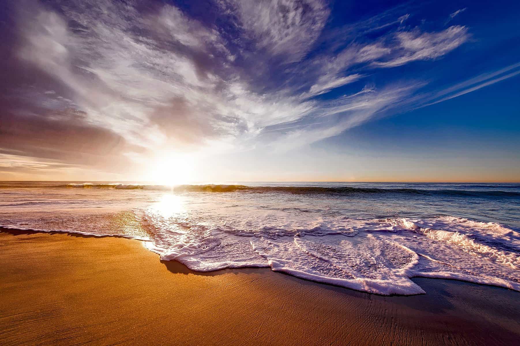 The sun is shown setting over the ocean by a camera held very low and waves are rolling over a sandy beach.
